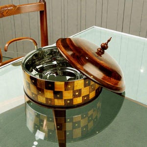 Puede incluir: Un cuenco decorativo para servir con tapa de madera. El exterior del cuenco presenta un patrón de tablero de ajedrez de cuadrados de madera clara y oscura, con pequeños detalles de estrellas. La tapa es de madera oscura con un remate decorativo. El interior es de acero inoxidable.