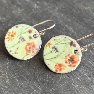 May include: A pair of round earrings with a floral design. The earrings feature a light green background with orange and red flowers, and small purple berries. The earrings have silver-colored ear wires.