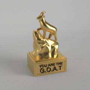 May include: A gold-colored trophy featuring a goat standing on a rock formation. The base of the trophy has the text "YOU ARE THE G.O.A.T." in black lettering. The trophy is set against a neutral background.