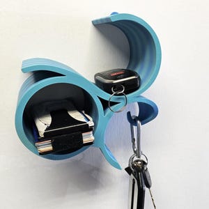 May include: A light blue, wall-mounted key and wallet holder. The holder has a unique, wave-like design with a shelf for a wallet and a key fob, plus a hook for keys. The holder is mounted on a white wall.