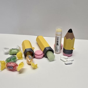 May include: Two yellow pencil-shaped plastic containers with gray accents and pink caps, one containing a lip balm tube. Three Jolly Rancher candies in green and red wrappers are nearby, along with four white oval pills and a lip balm tube.