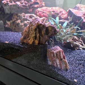 May include: A close-up of an aquarium with decorative rocks and gravel. The rocks are brown and reddish-brown, with a textured surface. The gravel is black. The aquarium is filled with water and has some plants in the background.