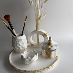 Puede incluir: Una bandeja decorativa blanca con un borde salpicado de oro contiene una colección de artículos de cerámica blanca. Estos incluyen un jarrón texturizado con pinceles, un jarrón en forma de anillo con flores secas y un pequeño cuenco en forma de loto, todos con detalles dorados.