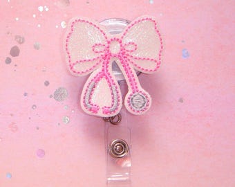 May include: White and pink glitter stethoscope bow badge reel. The bow is white with pink stitching and glitter. The stethoscope is silver with pink stitching. The reel is clear plastic with a silver button.