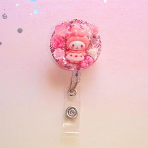 Könnte beinhalten: Ein pink-weißes Abzeichen mit einer Cartoon-Figur in der Mitte, umgeben von rosa Rosen und Glitzer. Das Abzeichen hat einen transparenten Kunststoffhalter und einen Metallclip.