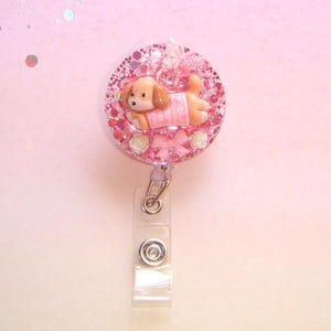 Könnte beinhalten: Ein pinkfarbenes Glitzer-Ausweis-Jojo mit einem hellbraunen Welpen, der einen rosa Pullover trägt. Die Rolle hat einen transparenten Kunststoffriemen und einen Metallclip. Dekorative Elemente sind rosa Rosen, eine Schleife und ein Schmetterling.