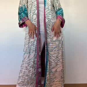 Peut inclure: Long kimono de couleur crème avec des détails turquoise et rose. Le motif floral orne la robe, avec des broderies florales turquoise sur les manches. Portée sur un pantalon blanc et un haut blanc, la robe est longue.