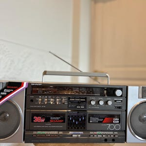 Peut inclure: Un radio-cassette Sharp gris avec un lecteur de cassette central, un tuner radio et des haut-parleurs détachables. L'appareil présente une puissance de 36W, un égaliseur graphique et une poignée de transport argentée. Texte Sharp, 36W, 700.
