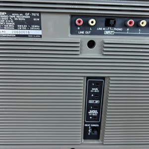 Pu&ograve; includere: Il pannello posteriore di un registratore radio stereo Sharp GF-767E grigio con lamelle di ventilazione orizzontali, un'etichetta con specifiche tecniche, porte RCA, terminali per altoparlanti esterni e un interruttore beat cancel.