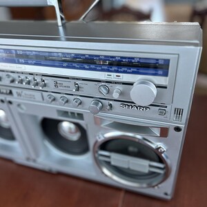 May include: A silver Sharp boombox featuring a prominent radio tuner display with blue accents, multiple circular speaker grilles, and various control knobs and switches. The front panel displays the Sharp brand name and technical audio settings.