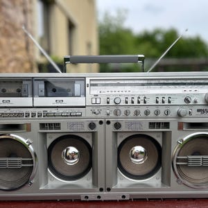 May include: A silver Sharp GF-777 boombox featuring dual cassette decks, a radio tuner with a frequency display, and four front-facing speakers. The portable audio system includes a top carrying handle and two extendable metal antennas.
