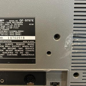 May include: A close-up of a grey Sharp GF-9797E stereo radio tape recorder. The back of the unit features a label with technical specifications, including frequency range and serial number 10702518. The unit has a textured surface and several holes.
