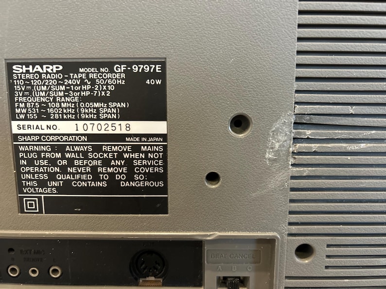 May include: A close-up of a grey Sharp GF-9797E stereo radio tape recorder. The back of the unit features a label with technical specifications, including frequency range and serial number 10702518. The unit has a textured surface and several holes.