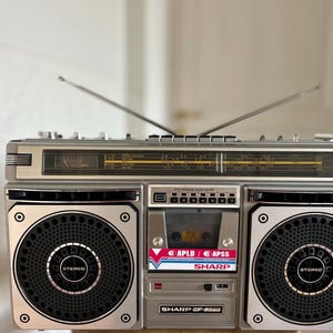 Könnte beinhalten: Ein silberner Sharp GF-8585 Stereo-Boombox mit zwei Lautsprechern, einem zentralen Kassettendeck mit APLD- und APSS-Beschriftung, einer Radiotuner-Anzeige mit gelben Frequenzmarkierungen und zwei ausziehbaren Metallantennen.