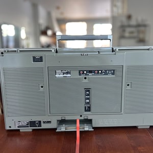 Pu&ograve; includere: Un boombox portatile grigio visto dal retro, dotato di una maniglia superiore per il trasporto, griglie per altoparlanti su entrambi i lati e un pannello centrale con varie porte di ingresso e uscita, inclusi jack RCA e un vano batterie.