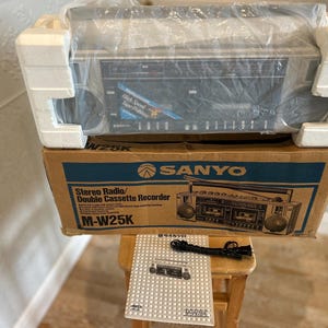 May include: A Sanyo M-W25K stereo radio and double cassette recorder in its original cardboard box with white polystyrene packaging. The box features blue text and a line drawing of the device. Includes a black power cord and a printed manual.