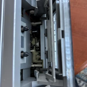 Pu&ograve; includere: Un lettore di cassette argentato con il meccanismo dello sportello aperto. L'interno rivela due perni neri, una testina di riproduzione magnetica centrale e componenti in plastica. Lo sportello in plastica trasparente &egrave; incernierato e inclinato.