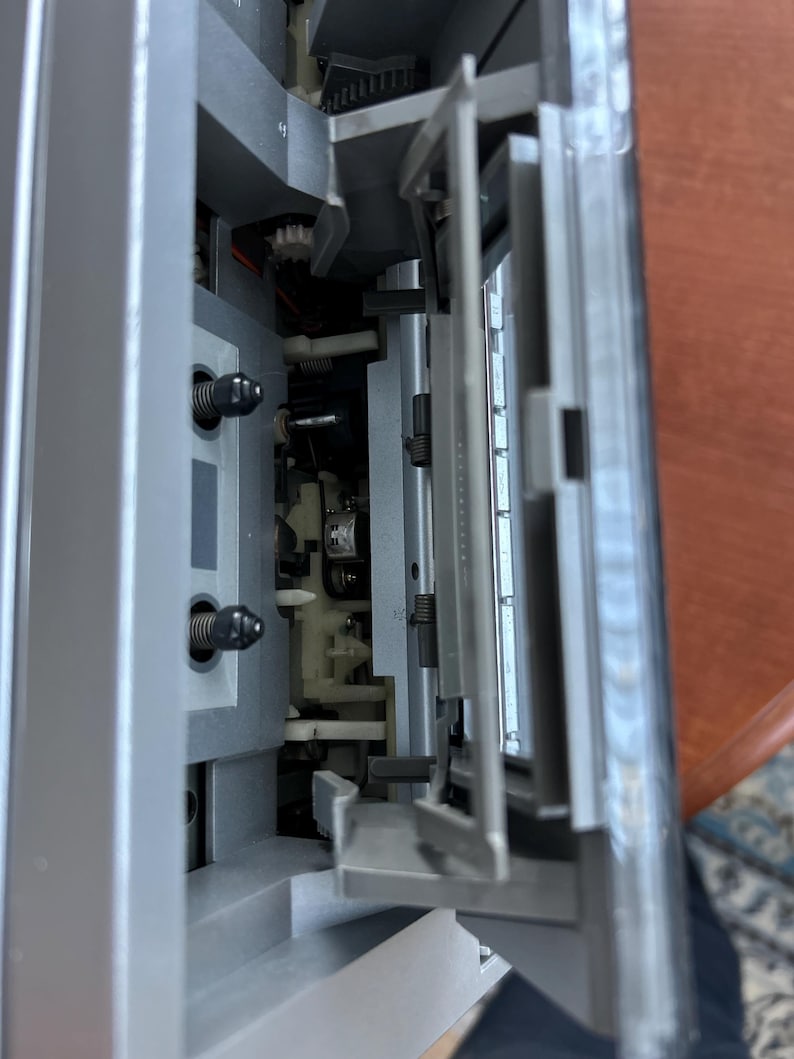 Pu&ograve; includere: Un lettore di cassette argentato con il meccanismo dello sportello aperto. L'interno rivela due perni neri, una testina di riproduzione magnetica centrale e componenti in plastica. Lo sportello in plastica trasparente &egrave; incernierato e inclinato.