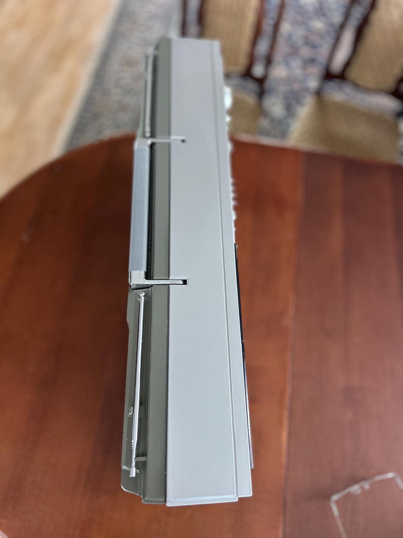Pu&ograve; includere: Un lettore radio-cassette portatile color argento e grigio visto di lato, dotato di un'antenna metallica retrattile, una robusta maniglia per il trasporto e un involucro rettangolare in plastica con comandi laterali e una griglia per altoparlante.