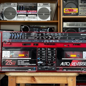 Puede incluir: Un radiocasete Sharp QT94 rojo y negro vintage con dos cubiertas de casete, ecualizador gráfico y función de inversión automática. El radiocasete tiene dos altavoces grandes y una salida de 25W. Otros equipos de audio vintage son visibles en el fondo.