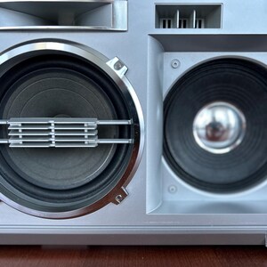 May include: A silver boombox speaker system featuring two large circular speakers with black cones. The left speaker has a horizontal silver grille overlay, while the right speaker features a central metallic dome. The housing is a matte silver finish.