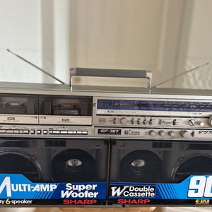 May include: A silver Sharp GF-787 portable stereo system with dual cassette decks, a radio tuner, and black speaker grilles. The front features blue and white labels reading Multi-Amp, Super Woofer, Double Cassette, and 90W PMPO with two antennas.