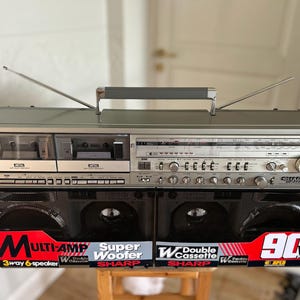 Puede incluir: Un radiocassette Sharp GF-777 plateado con dos pletinas de casete, un sintonizador de radio y dos grandes rejillas de altavoz negras. El frente muestra etiquetas rojas y blancas que dicen Multi-Amp, Super Woofer, Double Cassette y 90W.