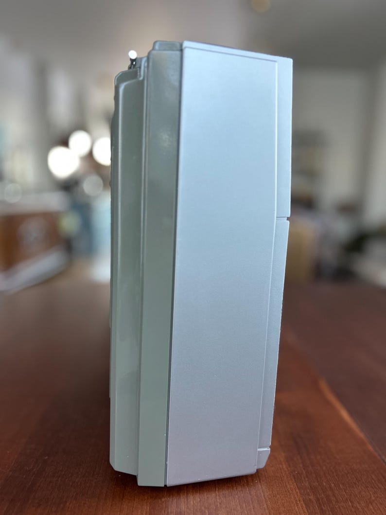 May include: A rectangular, silver-coloured electronic device with a smooth, matte finish. The side profile shows a vertical orientation with a slightly recessed panel and a small, metallic antenna tip protruding from the top edge of the casing.