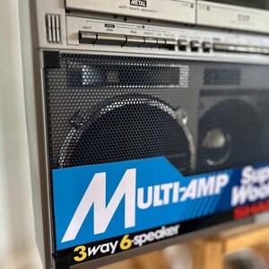 May include: A silver Sharp boombox featuring a black metal speaker grille and a blue label with white text that reads MULTI-AMP Super Woofer and 3way 6-speaker. The top section includes silver control buttons and a cassette deck labelled METAL.