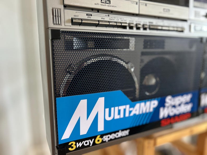 May include: A silver Sharp boombox featuring a black metal speaker grille and a blue label with white text that reads MULTI-AMP Super Woofer and 3way 6-speaker. The top section includes silver control buttons and a cassette deck labelled METAL.
