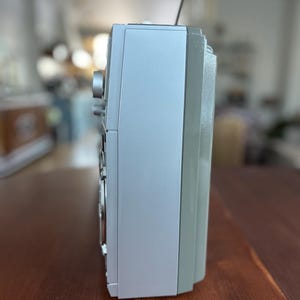 Pu&ograve; includere: Un profilo laterale di una radio portatile argentata con un involucro rettangolare liscio, una sottile antenna metallica che si estende verso l'alto dalla parte superiore e una manopola circolare visibile sulla parte anteriore del pannello.