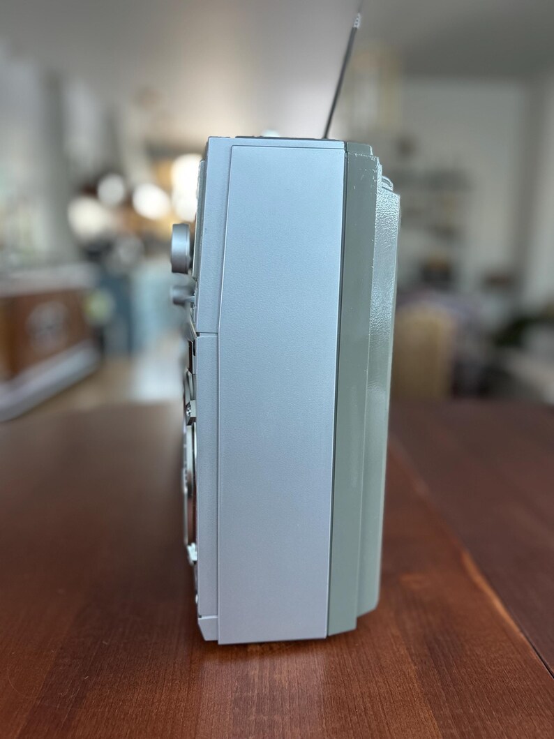 Pu&ograve; includere: Un profilo laterale di una radio portatile argentata con un involucro rettangolare liscio, una sottile antenna metallica che si estende verso l'alto dalla parte superiore e una manopola circolare visibile sulla parte anteriore del pannello.