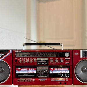May include: A red Sharp portable stereo system featuring dual cassette decks, a radio tuner, and detachable speakers. The unit displays text including Sharp, High Speed Dubbing, 700, APLD, and APSS. It includes a top handle and a metal antenna.