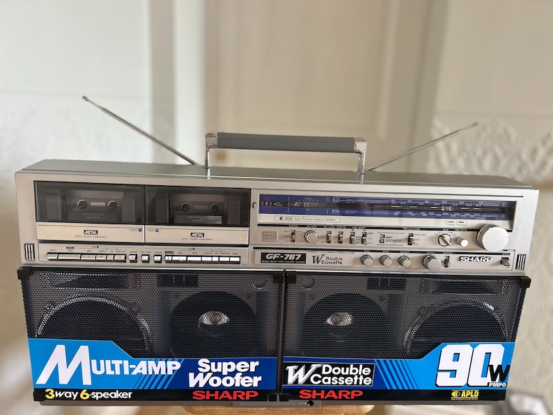 May include: A silver Sharp GF-787 portable stereo system with dual cassette decks, a radio tuner, and black speaker grilles. The front features blue and white labels reading Multi-Amp, Super Woofer, Double Cassette, and 90W PMPO with two antennas.