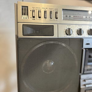 May include: A silver vintage boombox with a mesh speaker cover and various control buttons. The boombox features a tape selector, Dolby NR, function, and power buttons. The text on the boombox reads "tape selector", "Dolby NR", "function", and "power".