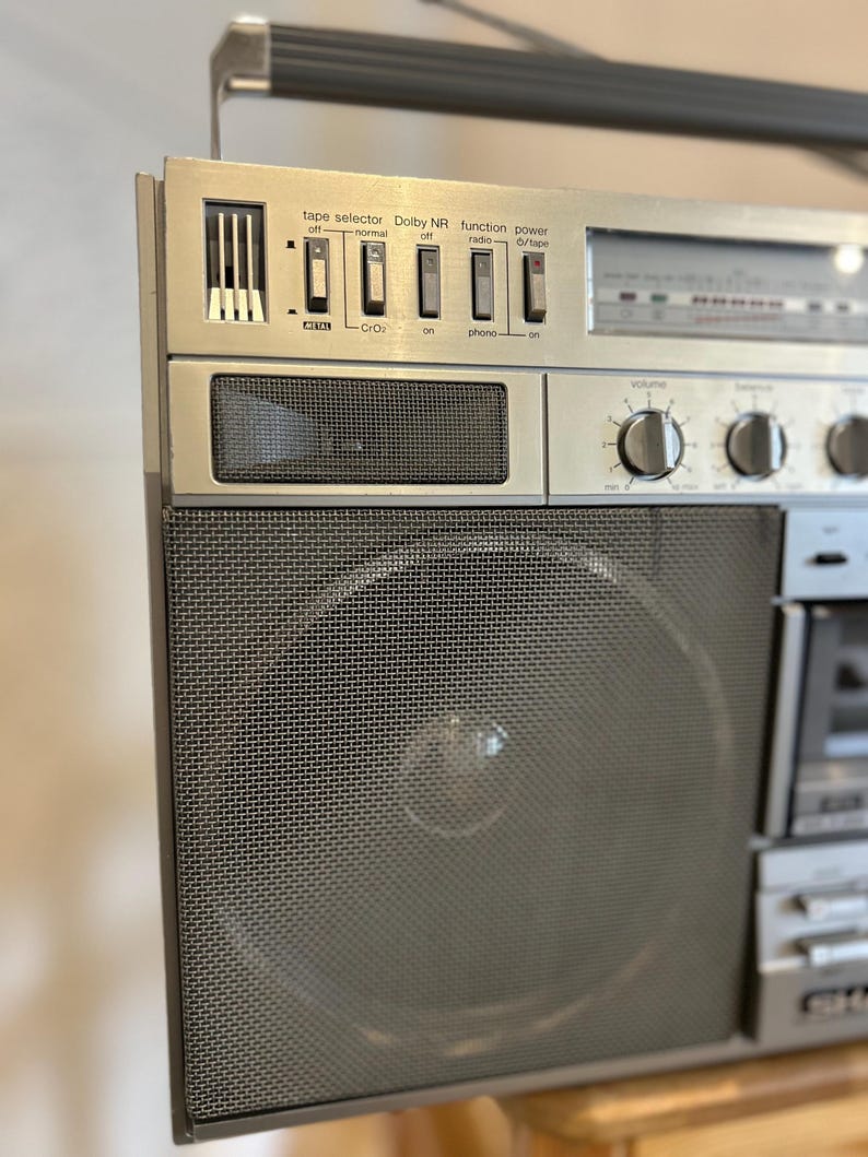 May include: A silver vintage boombox with a mesh speaker cover and various control buttons. The boombox features a tape selector, Dolby NR, function, and power buttons. The text on the boombox reads "tape selector", "Dolby NR", "function", and "power".