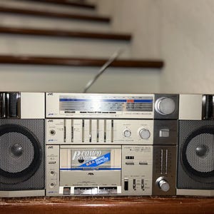 Op de afbeelding: Vintage JVC boombox stereo-ontvanger met luidsprekers. De zilver-zwarte stereo heeft een cassettespeler, grafische equalizer en radiotuner. De boombox heeft een intrekbare antenne en meerdere bedieningsknoppen.