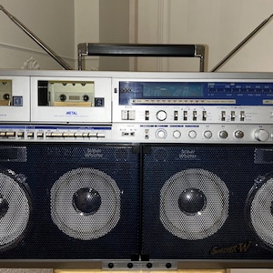 May include: Silver and black vintage boombox with dual cassette decks, a radio tuner, and large speakers. The boombox has a handle and two antennas. The brand name "SHARP" is visible on the front.