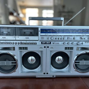 Pu&ograve; includere: Un radioregistratore a doppia cassetta Sharp GF-787 color argento con sintonizzatore radio e display blu, due vani per cassette, quattro altoparlanti frontali con dettagli metallici, maniglia superiore e antenna metallica estraibile.