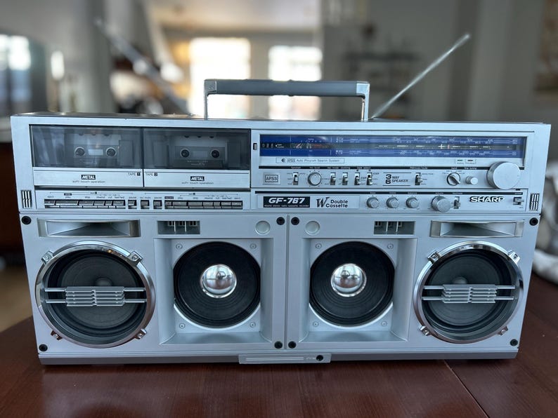 Pu&ograve; includere: Un radioregistratore a doppia cassetta Sharp GF-787 color argento con sintonizzatore radio e display blu, due vani per cassette, quattro altoparlanti frontali con dettagli metallici, maniglia superiore e antenna metallica estraibile.
