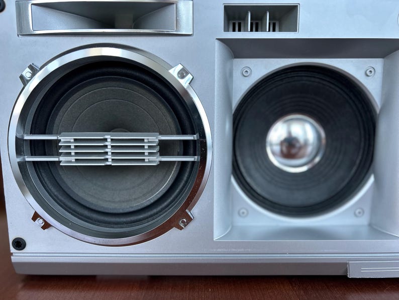 Pu&ograve; includere: Un sistema di altoparlanti boombox color argento con due altoparlanti circolari a cono nero. L'altoparlante sinistro presenta una griglia argentata orizzontale, mentre quello destro ha una cupola metallica centrale. L'unit&agrave; include prese d'aria rettangolari.