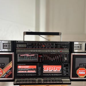 Peut inclure: Un radiocassette Sharp WF-939 noir doté de deux lecteurs de cassettes, d'un tuner radio et d'un analyseur de spectre. Les haut-parleurs portent la mention 200W et la marque Sharp. L'appareil comprend une poignée, des accents argentés et des boutons.
