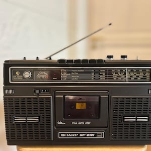 May include: A black Sharp GF-8181 portable radio cassette recorder featuring a top-mounted extendable antenna, a front-facing cassette deck with a clear window, dual speakers, and a radio frequency display panel with text including SHARP GF-8181.
