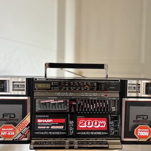 Puede incluir: Un radiocasete Sharp WF-939 negro con pletinas de casete dobles con auto-reverse, un analizador de espectro y un sintonizador de radio. Los altavoces indican una potencia de 200W y la unidad incluye un asa de transporte en la parte superior.