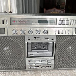 May include: Silver Sharp GF-9797 boombox with large speakers, a cassette player, and a radio dial. The boombox has multiple knobs and buttons, and the cassette player is visible in the center. The boombox is sitting on a patterned surface.