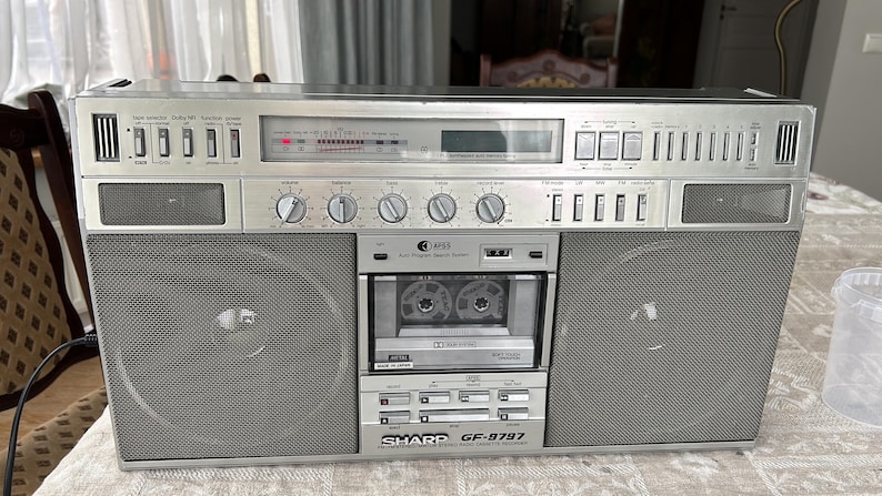 May include: Silver Sharp GF-9797 boombox with large speakers, a cassette player, and a radio dial. The boombox has multiple knobs and buttons, and the cassette player is visible in the centre. The boombox is on a patterned surface.