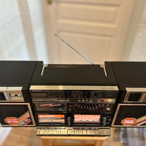 Puede incluir: Un radiocasete Sharp WF-939 negro con panel de control plateado, pletinas de casete dobles y altavoces extraíbles. La unidad presenta una etiqueta de 200W en las rejillas de los altavoces, una antena metálica retráctil y un sintonizador.