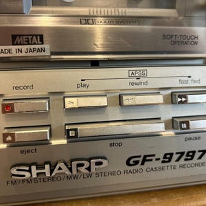 May include: Close-up of a silver Sharp GF-9797 stereo radio cassette recorder. Features include a cassette deck, control buttons for record, play, rewind, fast forward, stop, and pause. The text "METAL MADE IN JAPAN" is visible.