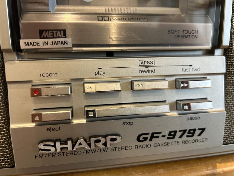 May include: Close-up of a silver Sharp GF-9797 stereo radio cassette recorder. Features include a cassette deck, control buttons for record, play, rewind, fast forward, stop, and pause. The text "METAL MADE IN JAPAN" is visible.