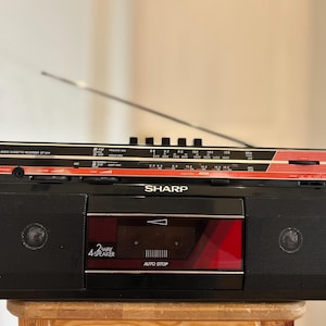 Puede incluir: Una grabadora de radio y casete estéreo Sharp de color negro con un panel de acento rojo, una antena telescópica plateada y un compartimento central para casetes. El frente presenta rejillas de altavoz negras, una etiqueta 4-way 2-speaker y un indicador auto stop.